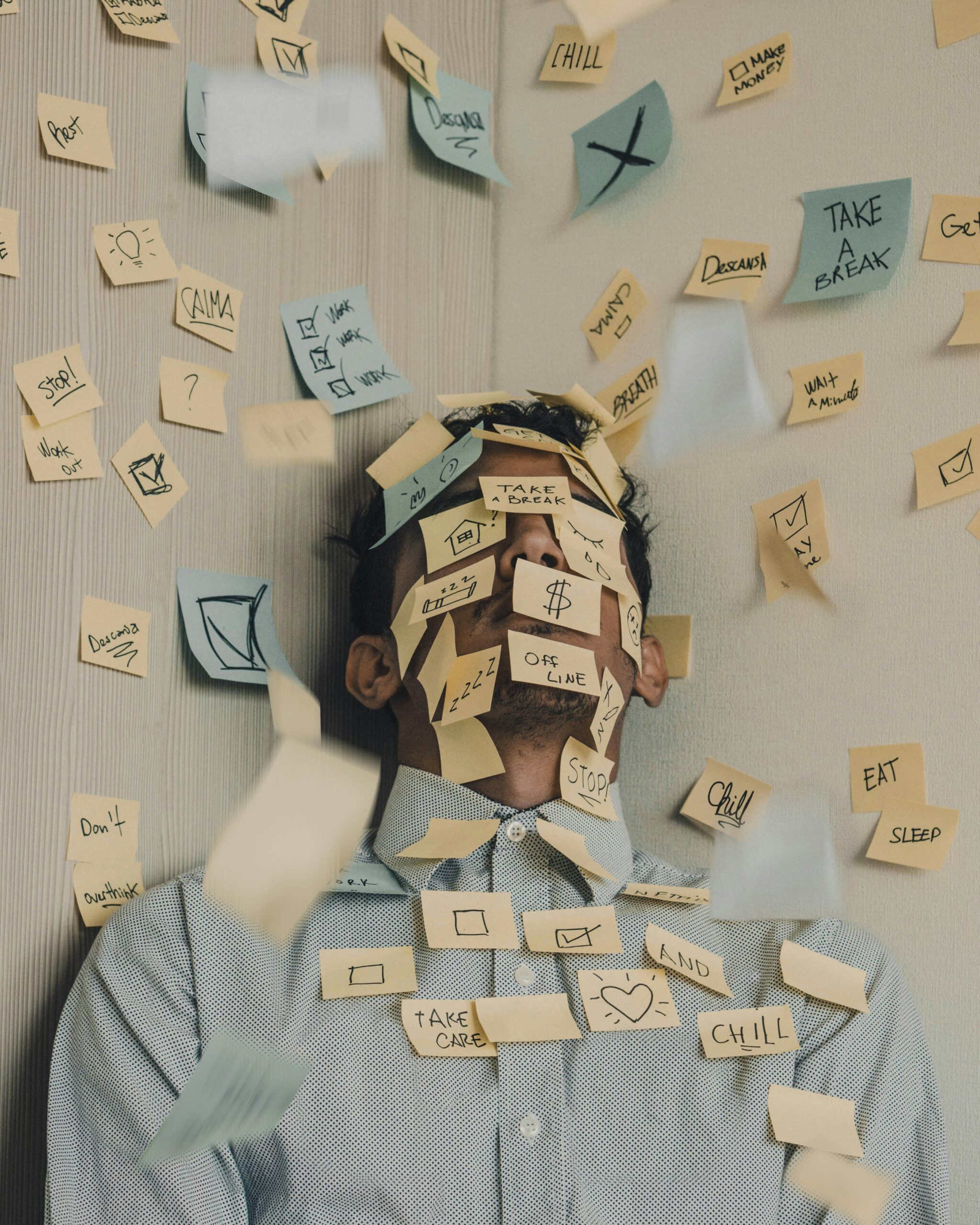 Person sitting against a wall with numerous sticky notes covering their face and falling around them, conveying a sense of overwhelm.