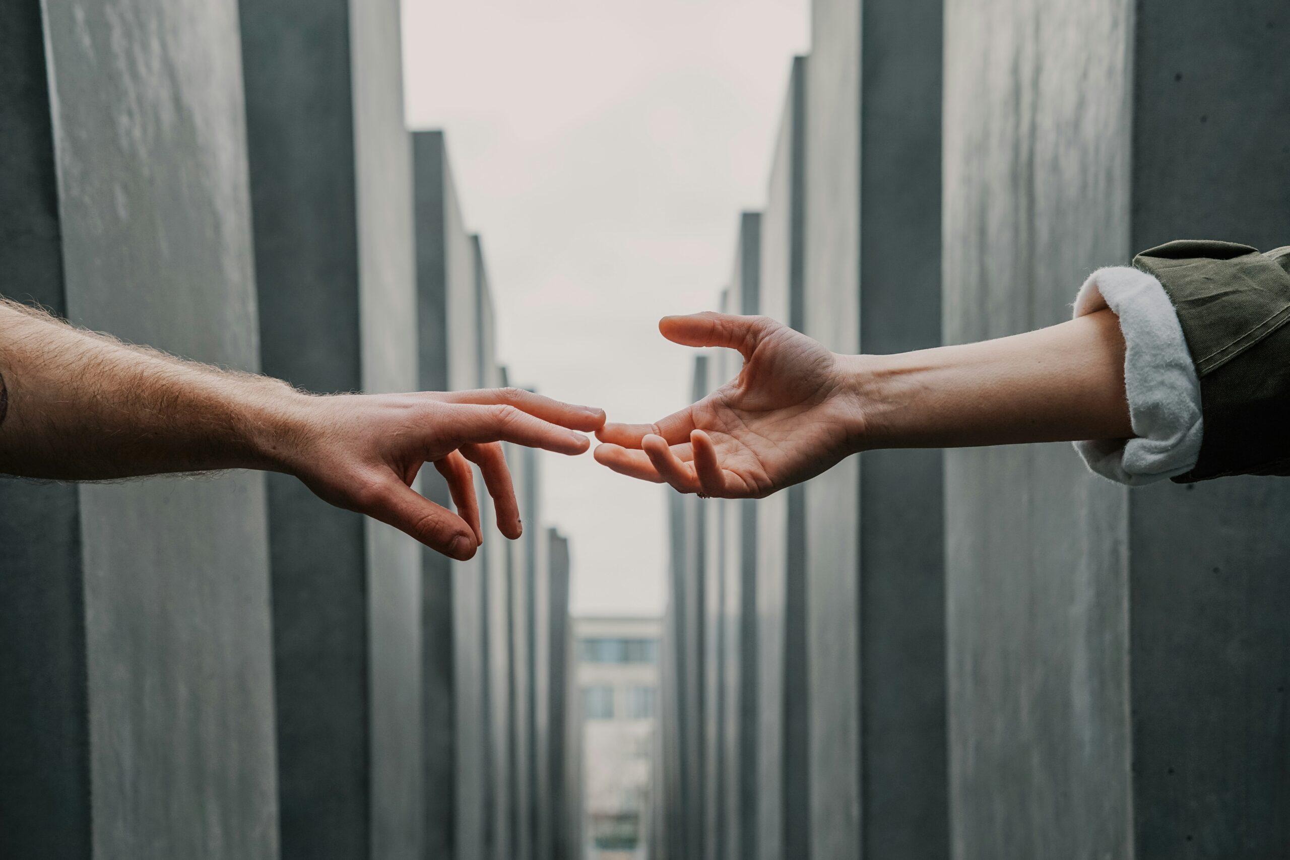 Two hands reaching toward each other with fingers nearly touching in a narrow, modern architectural corridor.