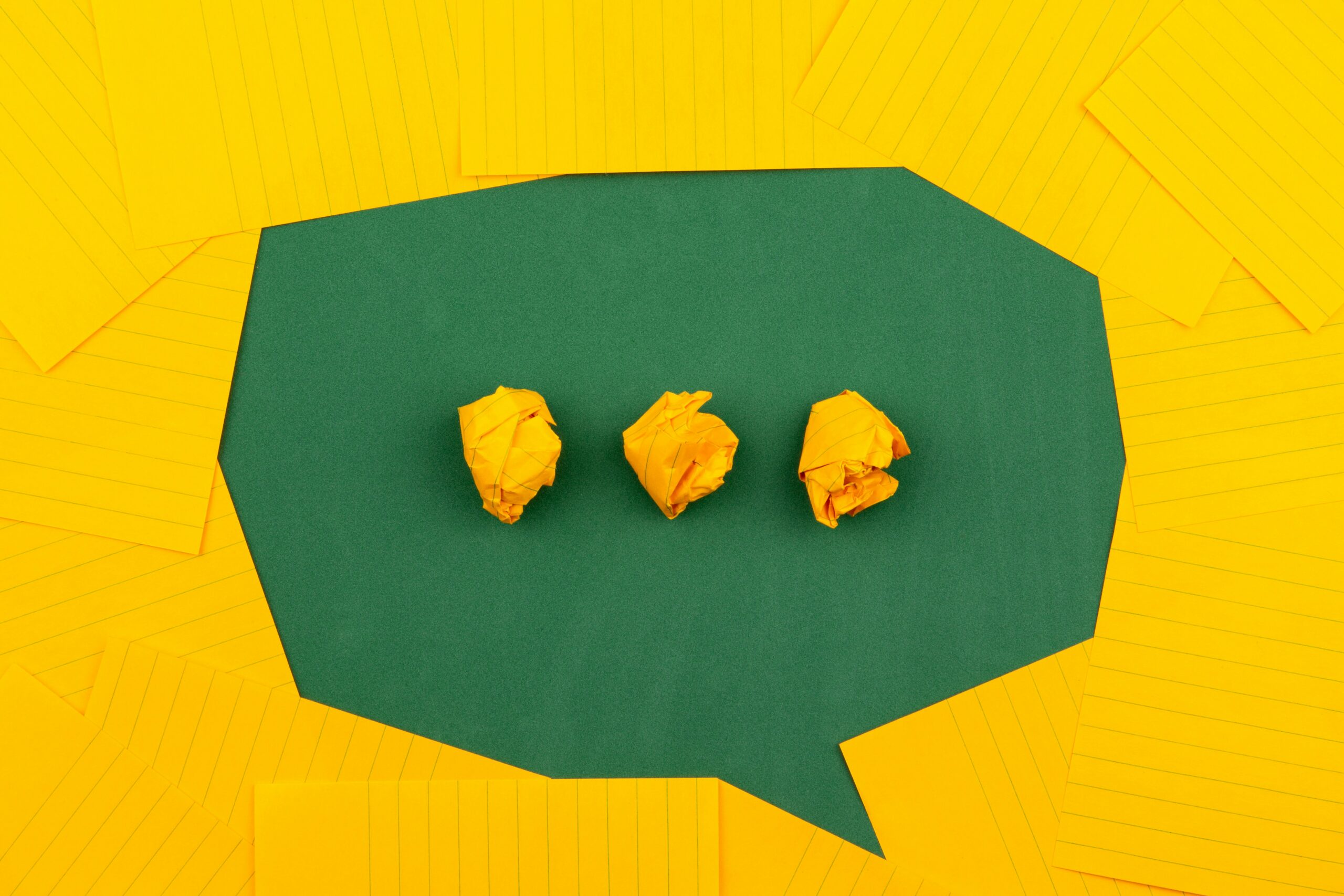 Three crumpled yellow paper balls on a green background resembling a speech bubble, surrounded by flat yellow sheets.