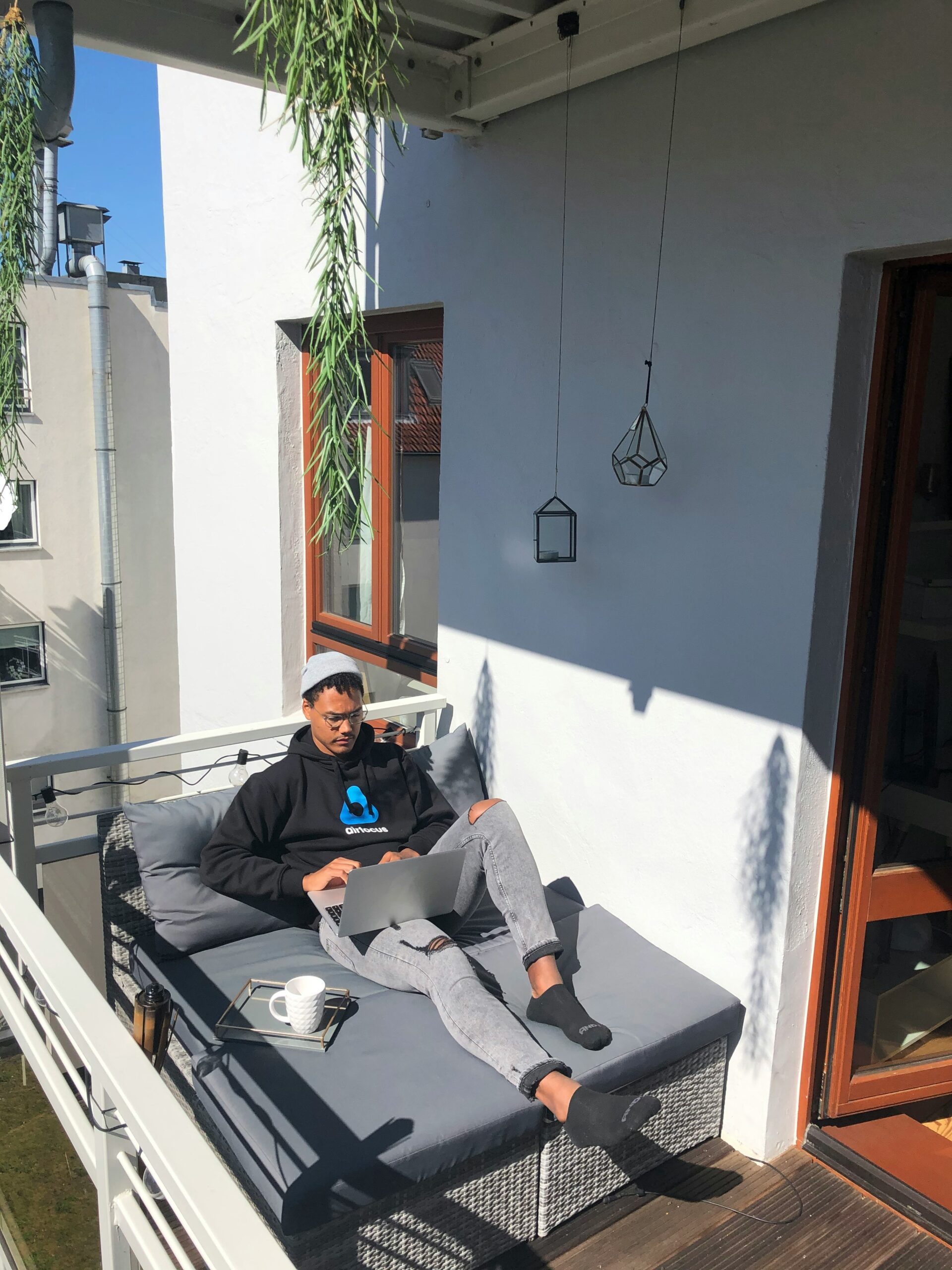 A person sits on a cushioned outdoor bench on a balcony, working on a laptop with a cup of coffee beside them. Sunlight casts shadows on the white wall and wooden floor.
