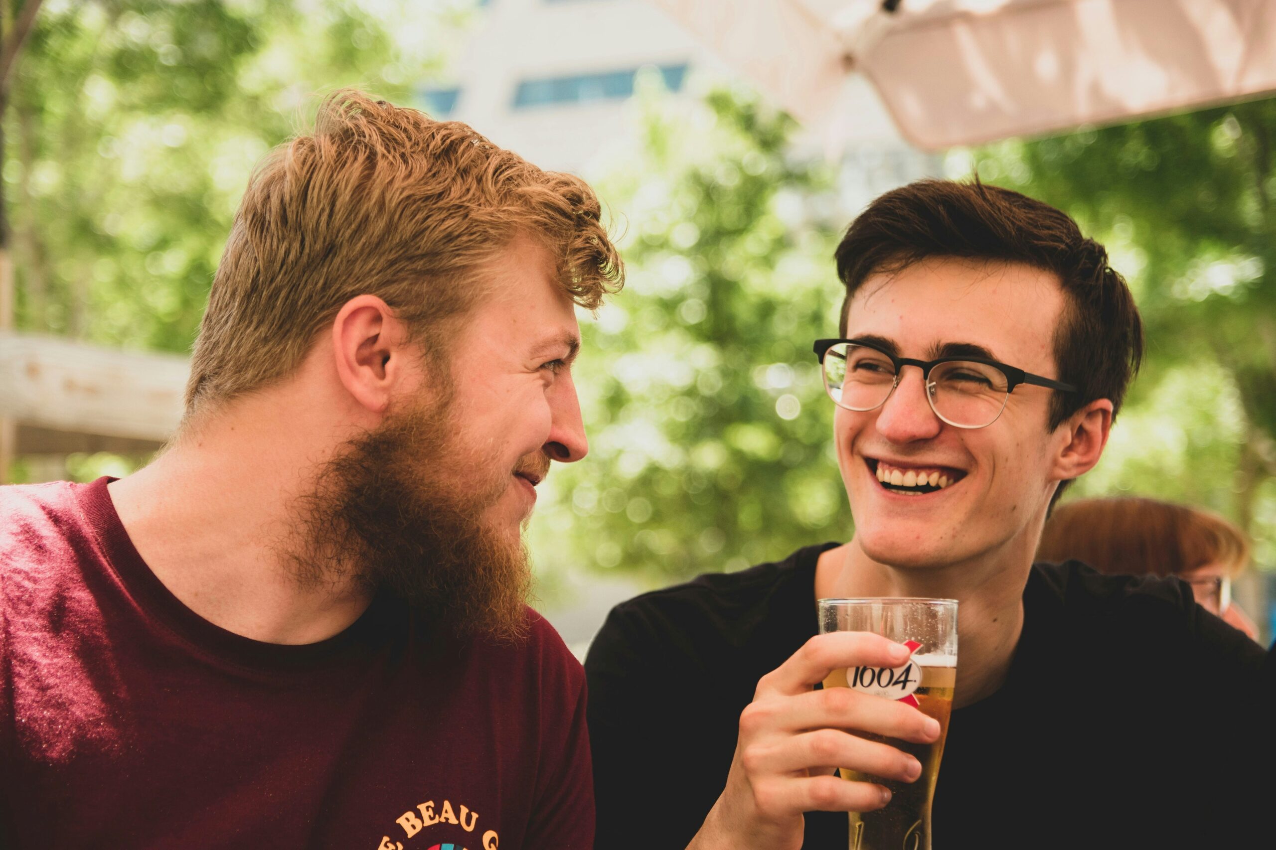 Two young men sit outdoors, one holding a glass of beer and smiling at the other. Both appear to be enjoying a casual conversation in a sunny setting.