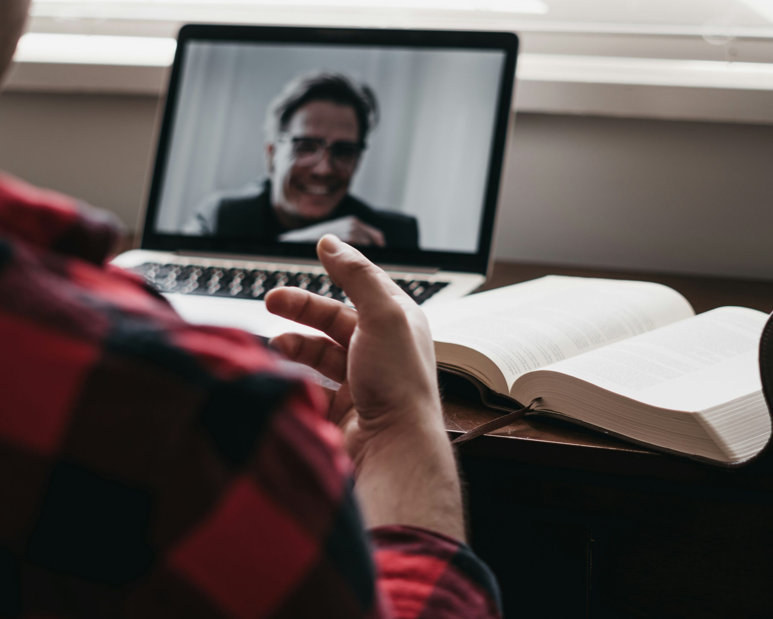A person in a red plaid shirt participates in a video call on a laptop, with an open book on the desk in front of them.