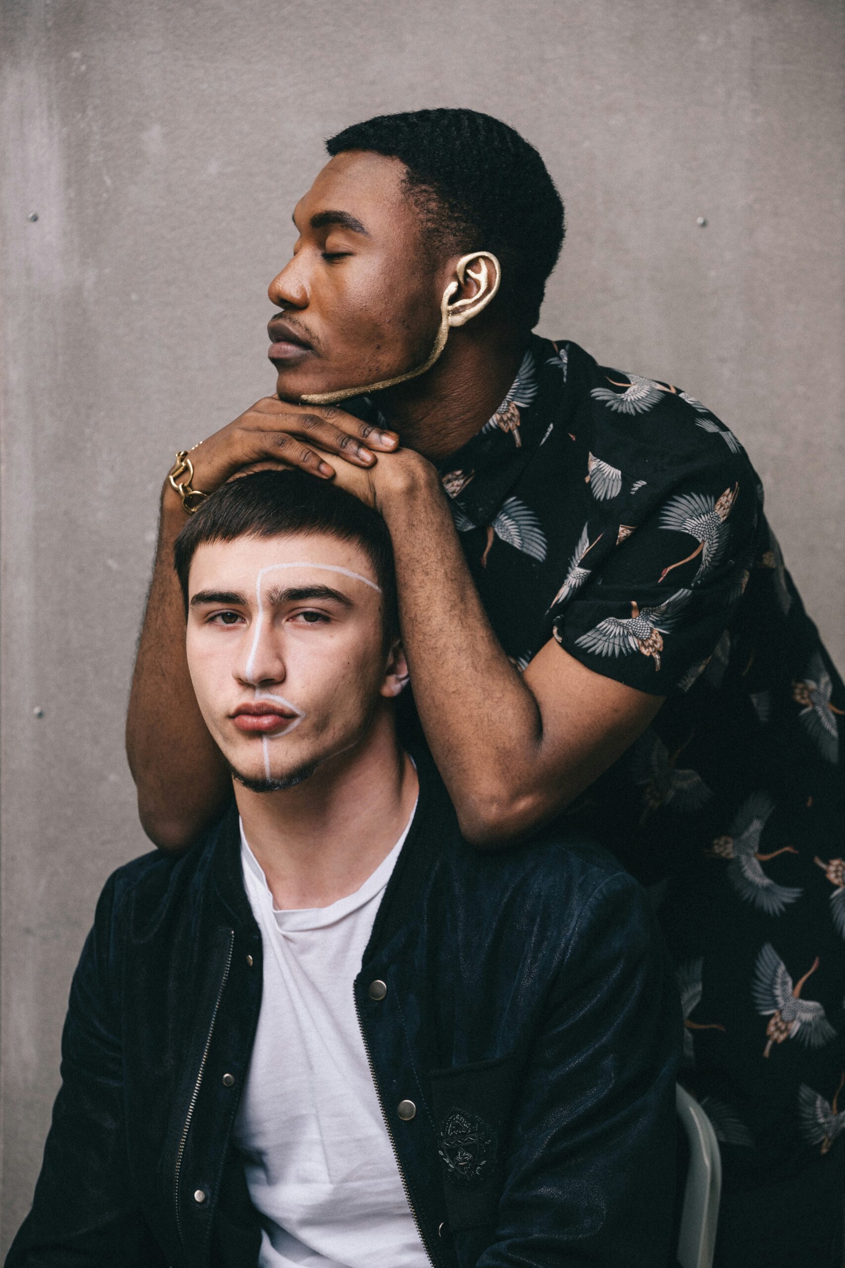 Two men pose against a plain background, one sitting with a neutral expression while the other stands behind, resting his arms on the seated man's shoulders. Both have painted lines on their faces.